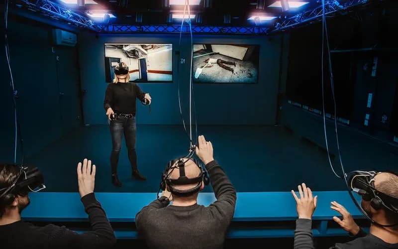Holodeck VR-Anwendung für das Bayerische Landeskriminalamt Eine Person, die ein VR-Headset trägt, interagiert in einem virtuellen Raum, der von drei anderen Personen mit Headsets beobachtet wird, in einem blau beleuchteten Raum mit Bildschirmen.