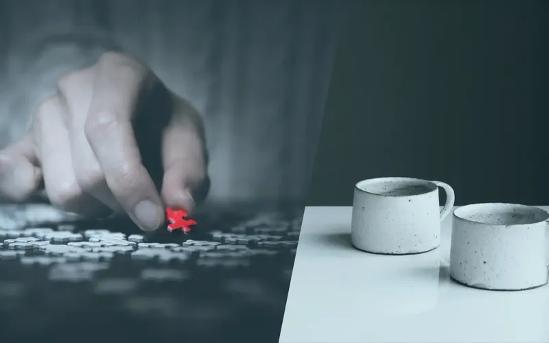 Kooperationspartner von INCAS Training - Headerbild Eine Hand legt ein rotes Puzzlestück auf ein schwarz-weißes Puzzlespiel. Zwei weiße Keramiktassen stehen auf einem Tisch rechts davon.